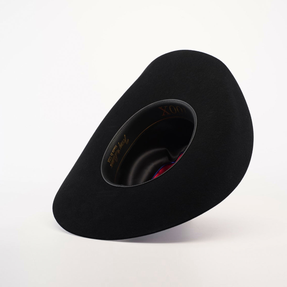 The 100X Tony Lama Black, a wide-brimmed hat made from premium beaver fur, rests on its side. It reveals an inner lining with a gold inscription and red ribbon detail, epitomizing genuine Western elegance against a plain white background.