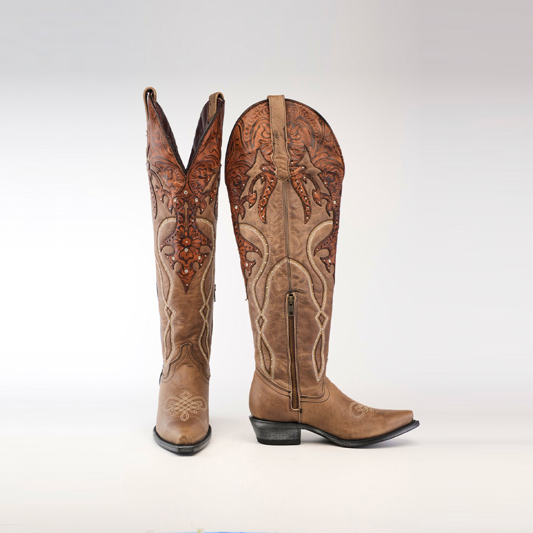 The Miriam Cappuccino Tall Shaft boots feature intricate embroidery and a tall decorative shaft, with one upright boot and the other leaning slightly, highlighting their handcrafted ornate patterns on the rich brown upper against a plain white background.