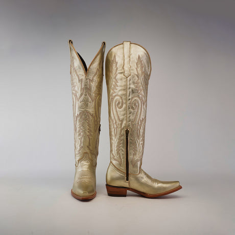 The Brittany Gold Tall Shaft Snip Toe boots feature intricate floral patterns on ornate leather, with one womens boot upright and the other showing a side zipper and pointed toe. Crafted with care, these gold treasures sit against a plain gray background with low wooden heels.