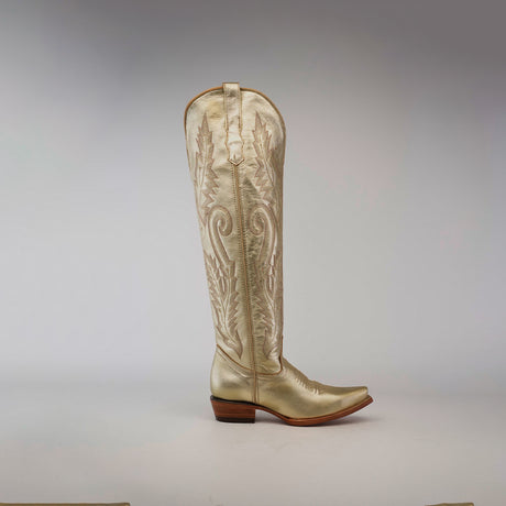 The Brittany Gold - Tall Shaft - Snip Toe womens boot is a metallic gold handcrafted masterpiece featuring intricate stitching and a low wooden heel. Set against a plain gray background, its pointed toe and embroidered shaft pattern make it a standout piece.