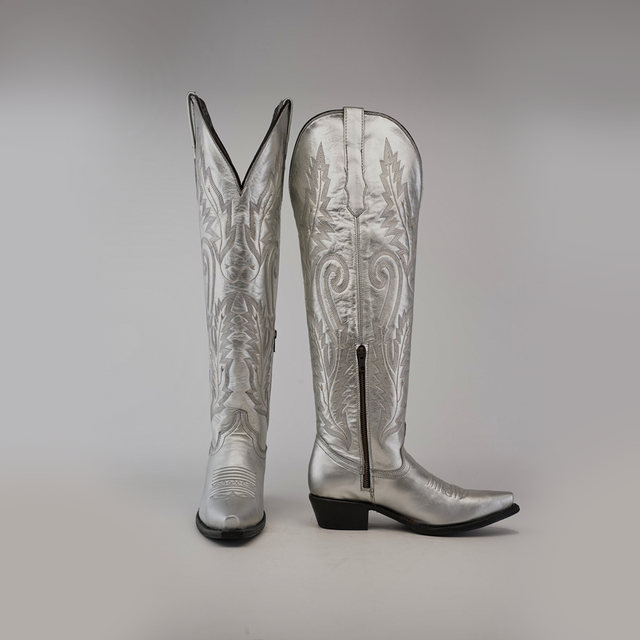 A pair of Brittany Silver Tall Shaft Snip Toe Boots, expertly crafted from premium leather with detailed stitching and side zippers. One boot stands upright while the other leans on a plain backdrop, highlighting their handcrafted elegance.