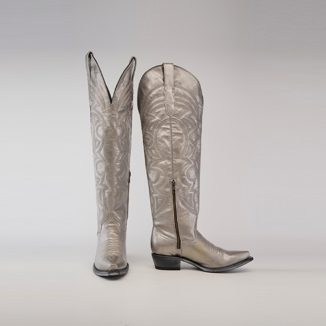 Two Taylor Mercury - Tall Shaft - Snip Toe boots, made of shiny metallic material with intricate swirling patterns. One stands upright, and the other is angled to show a side zipper and leather sole, set against a light gradient background.