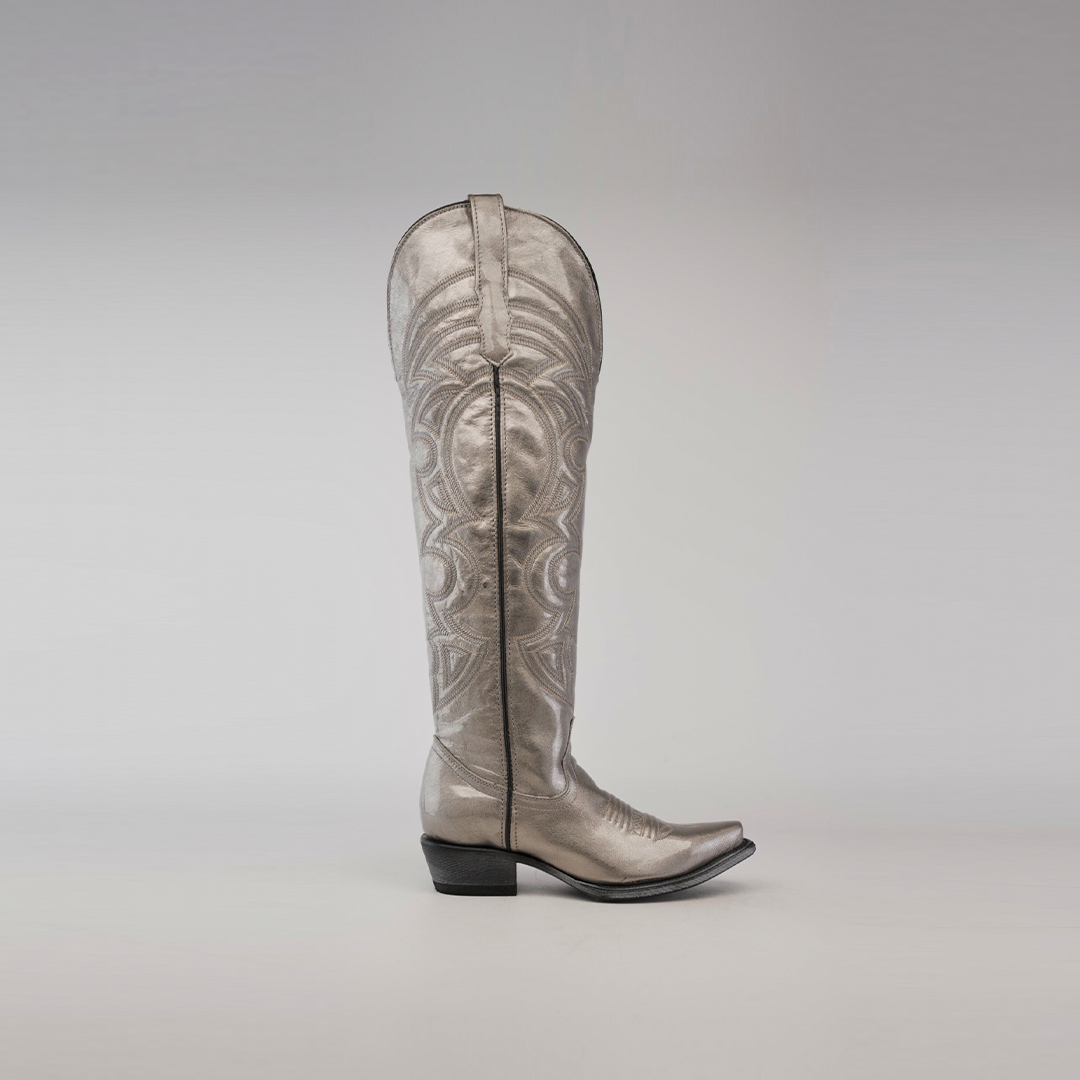 Against a plain gray background, the Taylor Mercury - Tall Shaft - Snip Toe cowboy boot stands out with intricate swirling patterns, a metallic silver finish, and a leather sole, showcasing elegance and craftsmanship.