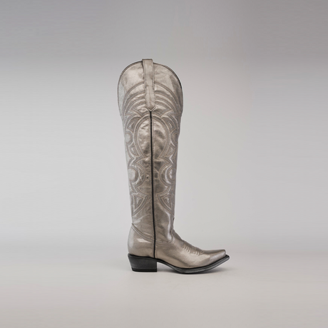 Against a plain gray background, the Taylor Mercury - Tall Shaft - Snip Toe cowboy boot stands out with intricate swirling patterns, a metallic silver finish, and a leather sole, showcasing elegance and craftsmanship.