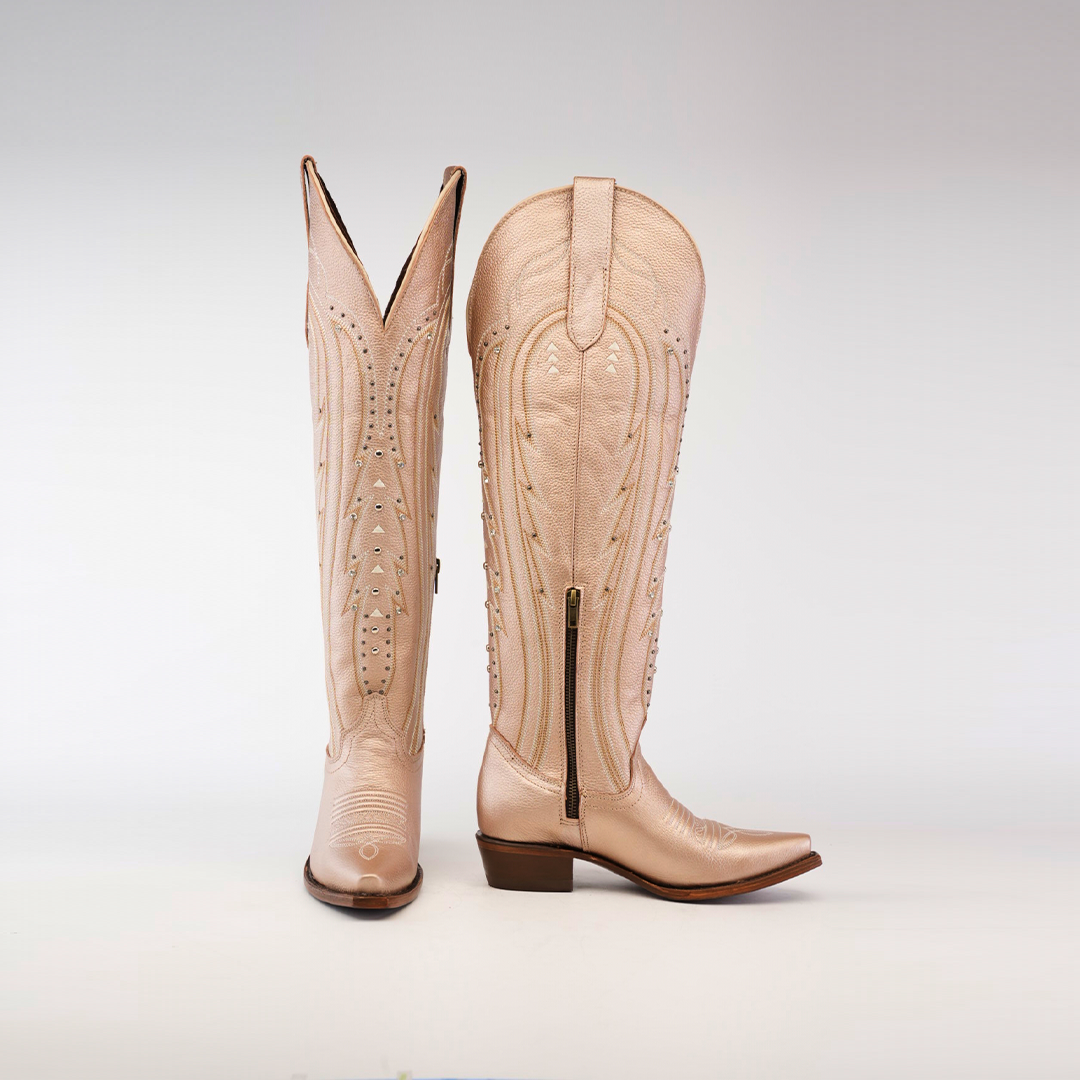 The Sara Metalic Florentine boots are tall beige cowboy boots with intricate stitching, featuring a snip toe, low heel, and visible side zipper. One boot is upright showing its front design while the other displays a side profile.