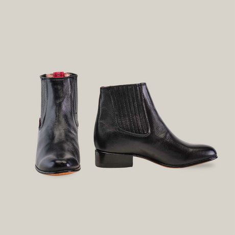 Against a light background, the Napa Black - Round Toe boots are displayed: one shows the front round toe, and the other highlights side elasticated panels for easy slip-on. Handcrafted with black Napa leather, they feature a slight heel and sleek design.