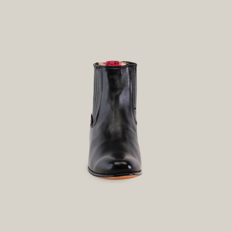 A front view of the Napa Black - Round Toe ankle boot in glossy black Napa leather shows a red pull tab on the back, with cowboy-inspired design. Its set on a light backdrop, casting a subtle shadow.