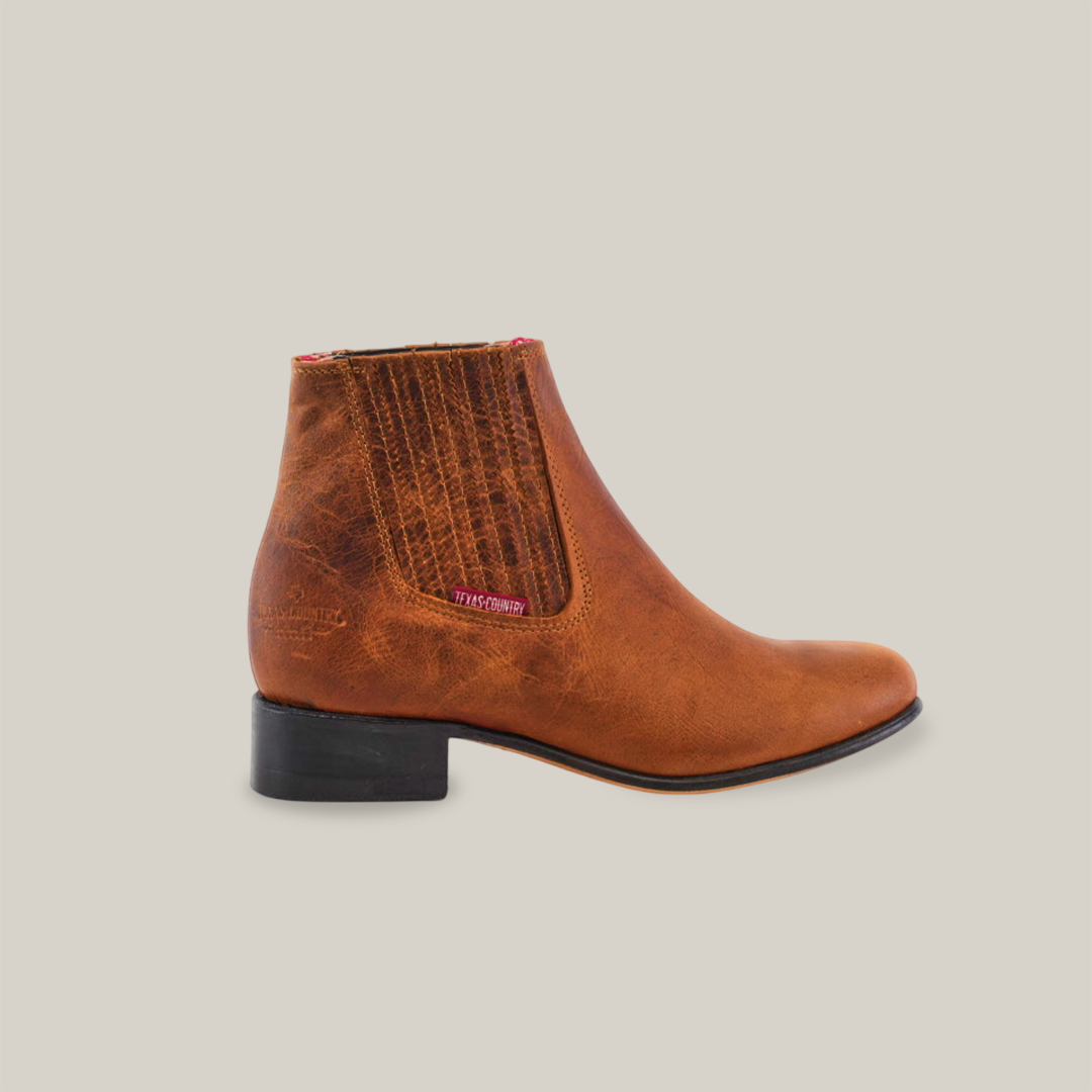 The Avejentado Honey - Round Toe boot in brown leather features a subtle sheen, low black heel reminiscent of classic western design, and pull-on style with stitched detailing and side fabric tag, all highlighted against a light background.