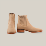 The Prime Suede Pearl Square Toe boots are handcrafted in beige suede with a wooden heel. The left boot faces forward, the right showcases the elastic side panel, all against a plain white backdrop.