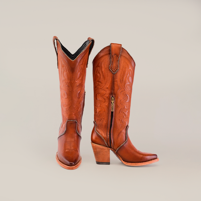 The Linda Tall Shaft Tan - J Toe, from the Platinum Collection, showcases premium leather brown cowboy boots with intricate stitching, pointed toes, and stacked heels against a plain light background. One boot stands upright while the other leans slightly, unveiling a gold zipper.