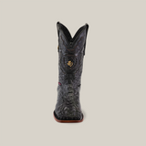 A black square-toed cowboy boot made from authentic exotic alligator leather, featuring intricate patterns and a striking decorative emblem on the front, elegantly displayed against a plain beige background.