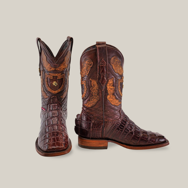 The Exotic American Alligator Hornback boots in brown showcase exceptional craftsmanship with intricately carved designs and a mix of crocodile textures. One boot stands upright, highlighting detailed patterns, while the angled one emphasizes its square toe design.