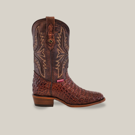 The Exotic Caiman Hornback - Brown - Round Toe is a unique handcrafted cowboy boot with intricate stitching on the shaft, featuring a textured reddish-brown caiman leather resembling reptile skin, a wooden heel, and a small red logo tag near the ankle.