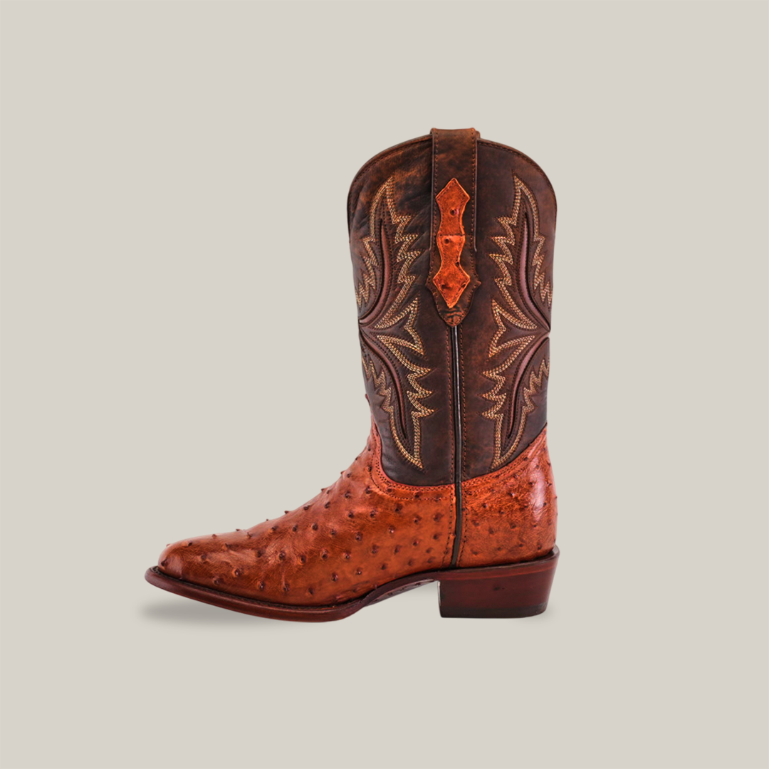 The Exotic Full Quilt Ostrich - Brandy - Round Toe is a single cowboy boot with a brown leather upper featuring decorative stitching and a foot and lower shaft made from textured reddish-brown ostrich leather, offering a striking two-tone handcrafted design.