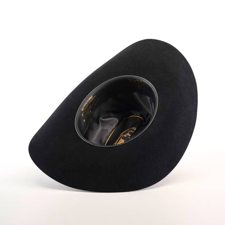 The 5X Dorado Black hat from Milano Hat Co., crafted from wild rabbit and beaver fur felt, rests upside down on a white background. It features a smooth, curved shape with glossy inner lining and gold text near the rim, embodying the elegance of a timeless western headpiece.