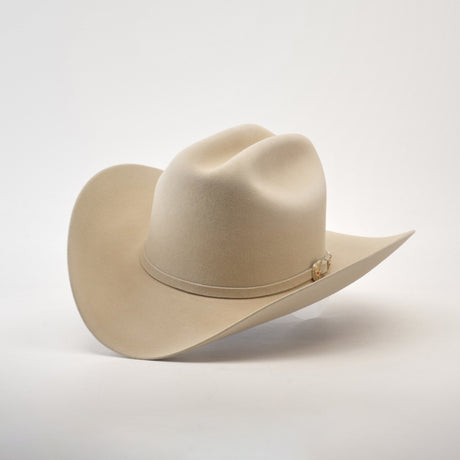 The 6X Real Belly, a light beige fur felt hat with a wide brim and decorative band, rests on a smooth white surface. Its premium Western design features a high crown with two slight dents that enhance its classic style.