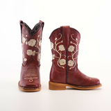 A pair of red cowboy boots, akin to Jatziry Cogñac - Square Toe, is shown on a white background. Adorned with intricate white floral embroidery and wooden soles, one boot is upright while the other leans to the side—ideal for stylish little girls.