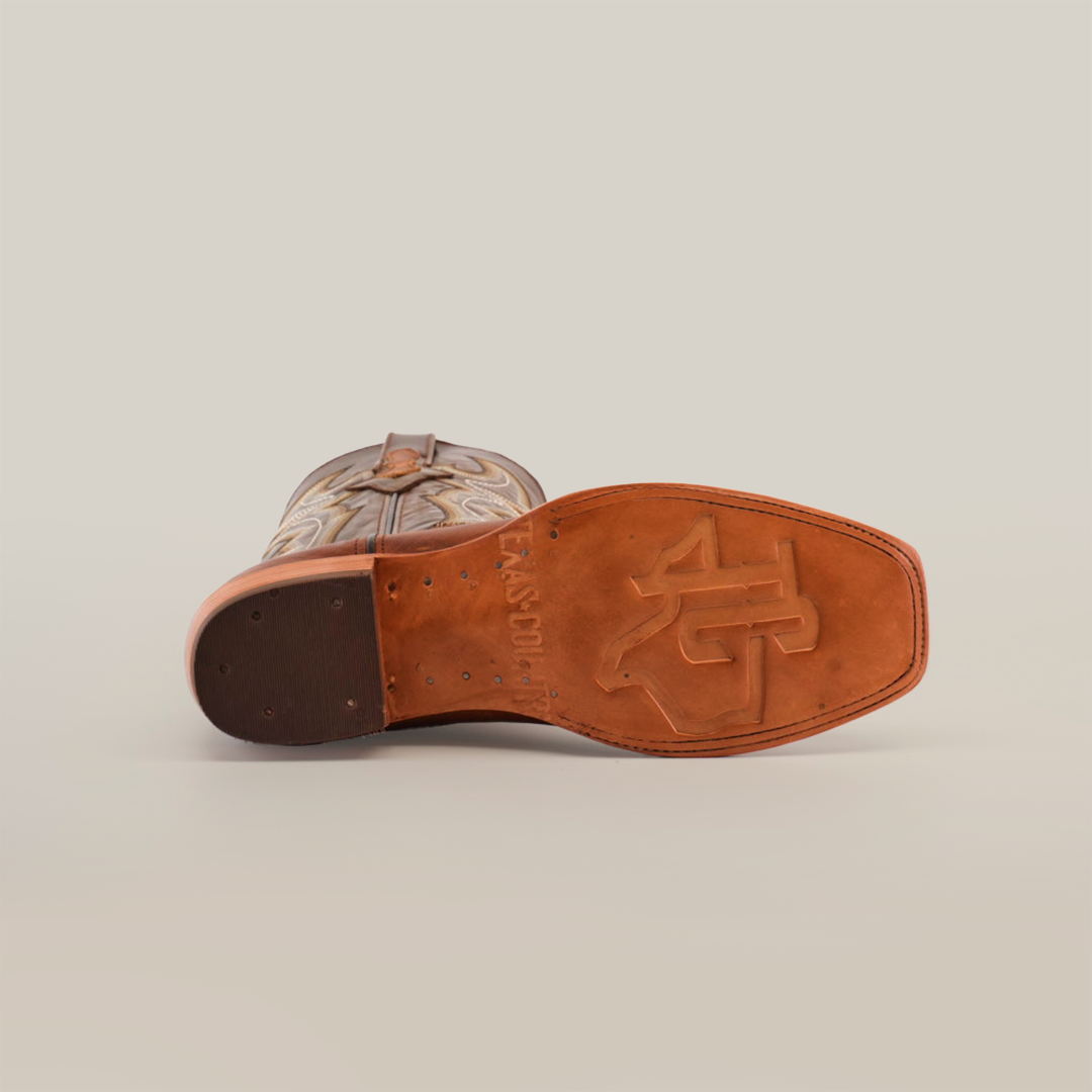 A close-up of the Fresno Tan - Wide Cutter Toe cowboy boot sole reveals intricate leatherwork that embodies the cowboy lifestyle. Made from premium cowhide leather, the tan sole has an imprinted logo, with decorative stitching on the upper part against a white background.