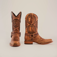 The luxurious Siberia Fawn - Biker Toe cowboy boots are crafted from premium cowhide, featuring intricate floral patterns and decorative stitching. A sturdy heel and a strap with metal accents across the vamp enhance their elegance, set against a plain white background.