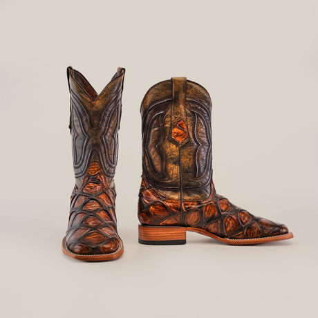 A pair of artisan boots crafted with rich Pirarucu Print Tan leather, high shaft, detailed stitching, wooden heel; one stands upright while the other lies sideways to display its intricate pattern. Product: Pirarucu Print Tan - Square Toe.