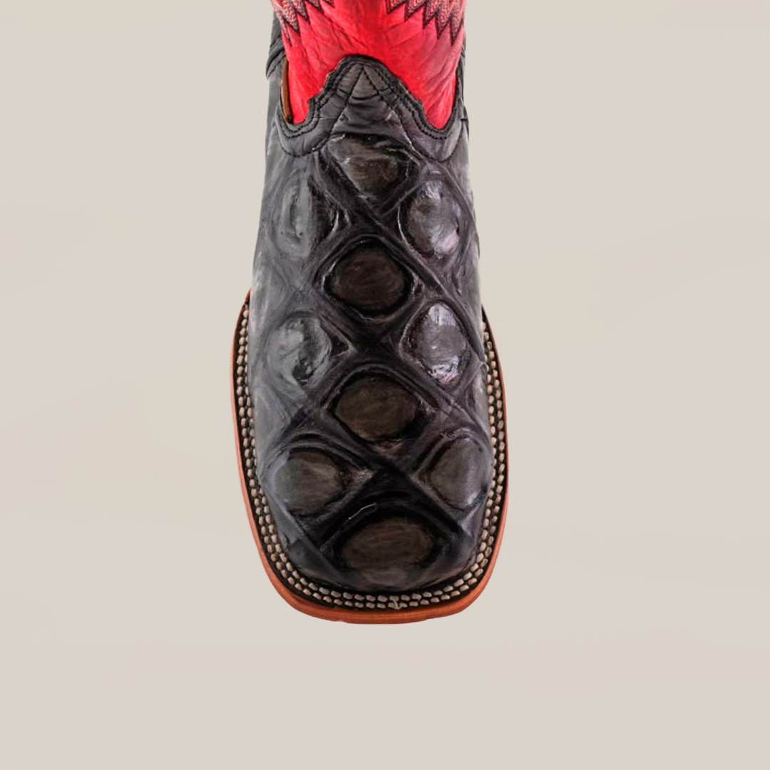 The Big Bass Pirarucu Print Rustic Black - Square Toe boot features a close-up of its black, textured premium cowhide leather toe with an exotic Pirarucu Print. Its decorative red upper contrasts elegantly with the sole outlined in crisp white stitching.