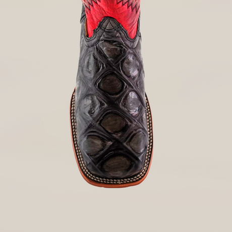 The Big Bass Pirarucu Print Rustic Black - Square Toe boot features a close-up of its black, textured premium cowhide leather toe with an exotic Pirarucu Print. Its decorative red upper contrasts elegantly with the sole outlined in crisp white stitching.