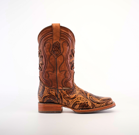 The Handtooled Leave Orix cowboy boot, made of premium brown cowhide leather, features intricate floral and swirl embossing on the shaft and foot. With a low wooden heel, rodeo toe, and slip-resistant sole, it combines style with functionality against a plain white backdrop.