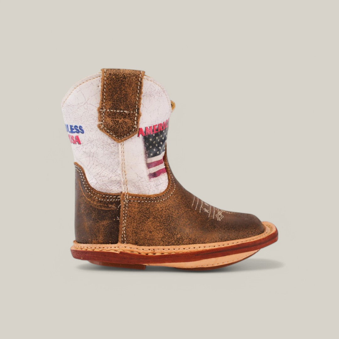 The Scott - Camel USA Shaft - Square Toe boot features a white upper with an American flag and AMERICALESS 1984 text. This trendy, premium footwear has a natural wood-colored sole and is set against a simple gray background.