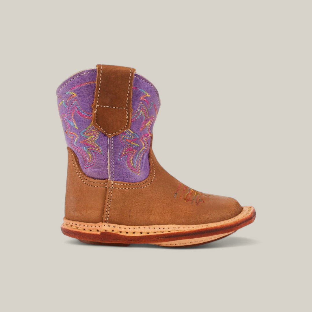 The Lilly boot, crafted in camel suede, features a distinctive violet upper shaft with colorful embroidery. It has a stylish square toe and visible stitching, standing out beautifully against a plain light beige background.