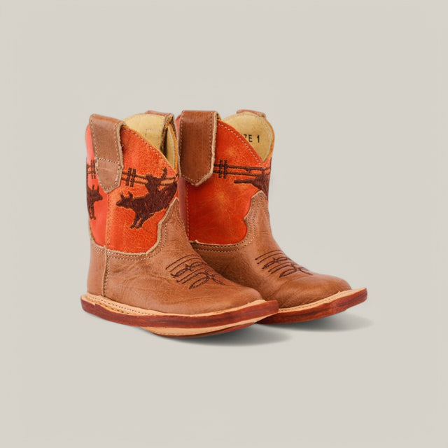 The Dillon Honey Bull baby boots feature an orange shaft with a rodeo rider silhouette on a brown leather body. They have square toes and slightly raised heels, set against a plain, light-colored background.