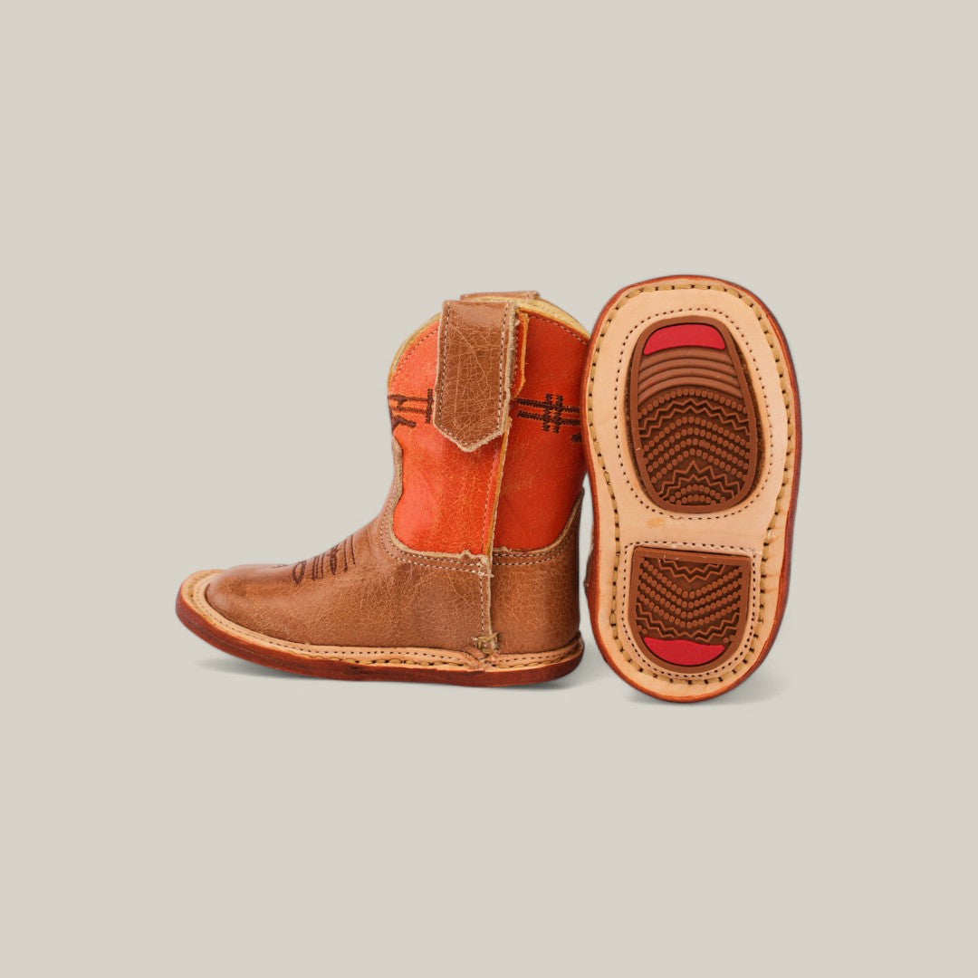 The Dillon - Honey Bull Orange Shaft - Square Toe is a charming pair of leather baby boots. One boot stands upright, featuring a vibrant orange shaft and brown upper, while the other lies on its side, revealing a textured sole with bold red accents, set against a plain light gray background.