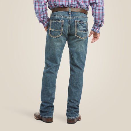 Wearing the M4 Low Rise Boundary Boot Cut Jean - Gulch with decorative stitching and paired with a plaid shirt, brown boots, and a belt, a person is comfortably viewed from behind against a soft beige background.