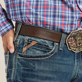 A person in a blue plaid shirt and relaxed fit M4 Low Rise Boundary Boot Cut Jeans in Gulch with a brown leather belt, showcasing a large Champion Header 2005 belt buckle. Their left hand rests near the pocket of the jeans.