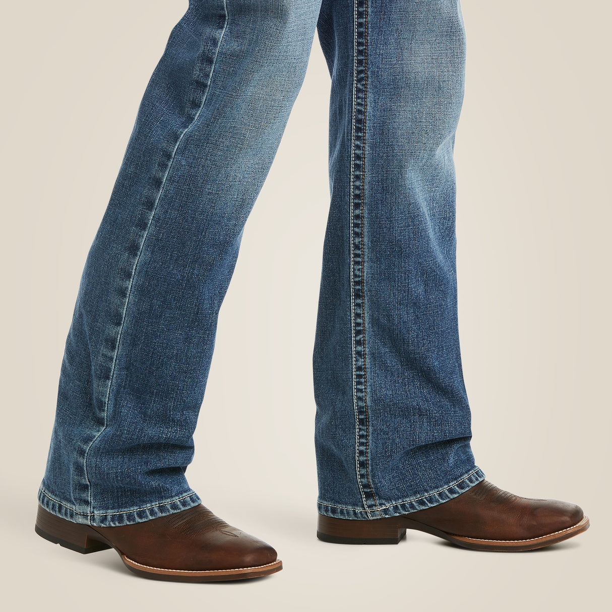 Close-up of someone wearing M5 Slim Stretch Stillwell Stackable Straight Leg Jean - Fargo over brown leather cowboy boots, slightly faded and elegantly draping over the boots against a plain, light background.