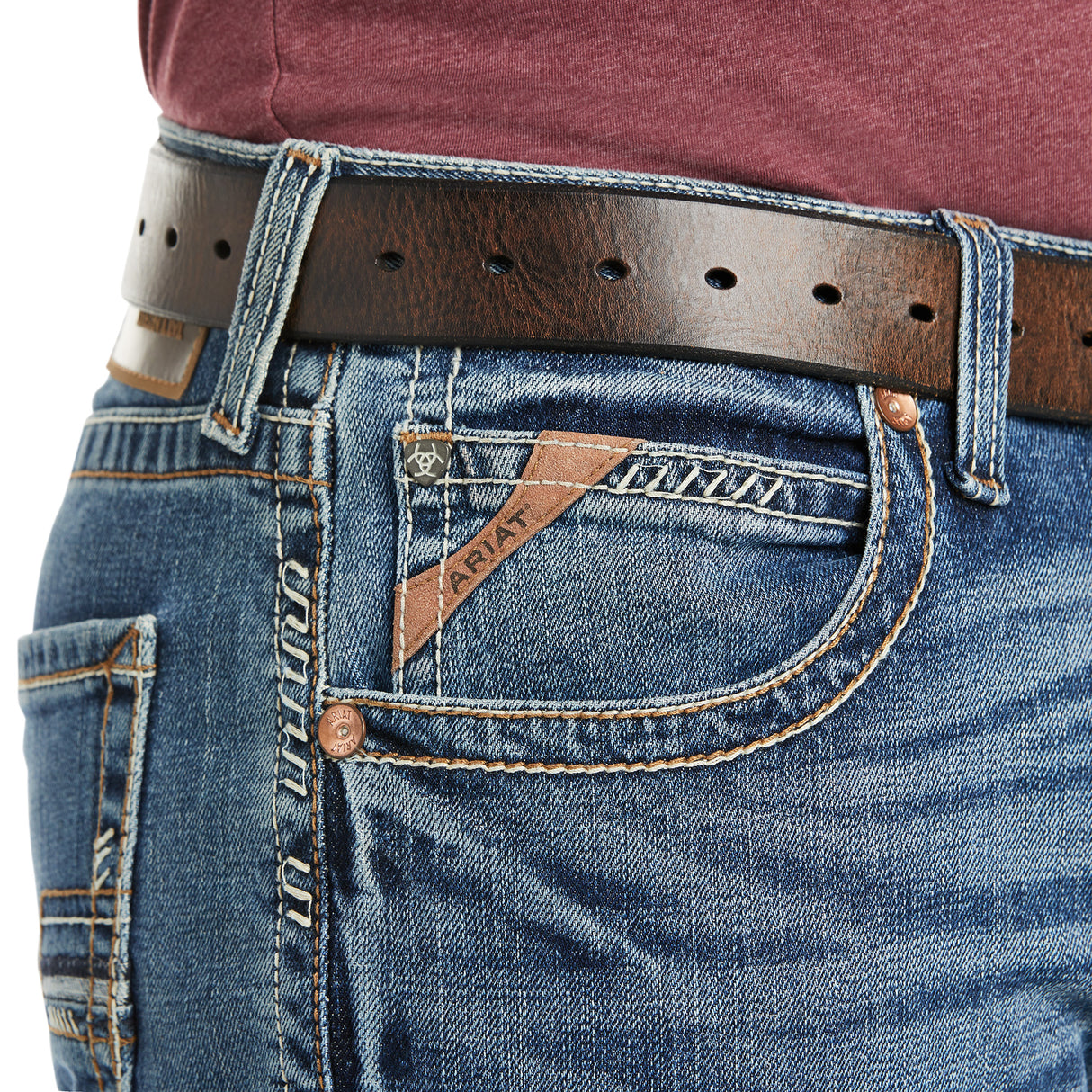 A close-up of someone wearing M5 Slim Stretch Stillwell Stackable Straight Leg Jeans - Fargo shows premium cotton blue denim with detailed stitching and a pocket logo. A brown leather belt with metal holes loops through the belt loops, effortlessly complementing the maroon shirt.