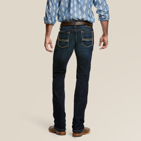 A person in a blue patterned shirt and dark jeans faces away, wearing Ariats M7 Rocker Concord Stretch Stackable Straight Leg Jeans in Dodge. These jeans feature prominent stitching and pockets, crafted for comfort with stretch fabric. The outfit is finished with brown shoes and a belt.