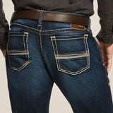 Shown from the back, a person wears M7 Rocker Concord Stretch Stackable Straight Leg Jeans in dark blue with beige stitching. The jeans feature stretch fabric, a straight leg fit, and two back pockets with decorative stitching. They also wear a black shirt and a brown leather belt.