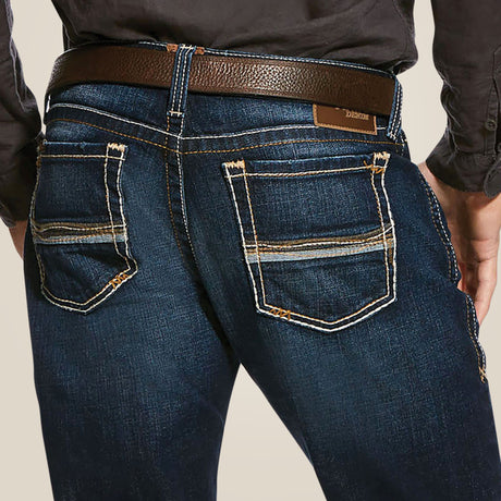 Shown from the back, a person wears M7 Rocker Concord Stretch Stackable Straight Leg Jeans in dark blue with beige stitching. The jeans feature stretch fabric, a straight leg fit, and two back pockets with decorative stitching. They also wear a black shirt and a brown leather belt.