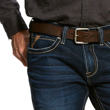 A person is wearing a dark long-sleeve shirt tucked into Ariats M7 Rocker Concord Stretch Stackable Straight Leg Jeans, secured with a brown leather belt. Their right hand rests casually in the front pocket of the stretch fabric jeans, focusing on the waist and hand.