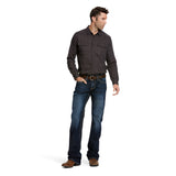 A man in a dark button-up shirt and M7 Rocker Concord Stretch Stackable Straight Leg Jeans stands on a white background, looking left. With one hand in his pocket, he complements his outfit with brown shoes.