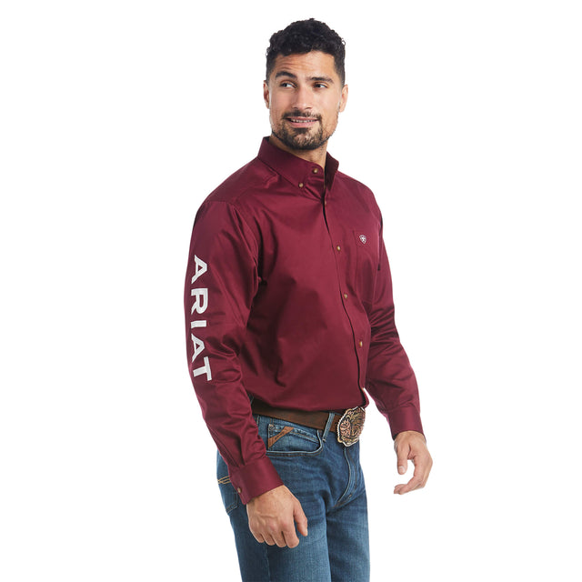 A man in a Team Logo Twill Classic Fit Shirt Burgundy - 10027995, featuring ARIAT on the right sleeve, blue jeans, and a decorative belt buckle, stands against a white background. His short dark hair and beard enhance his relaxed expression as he gazes to the side.