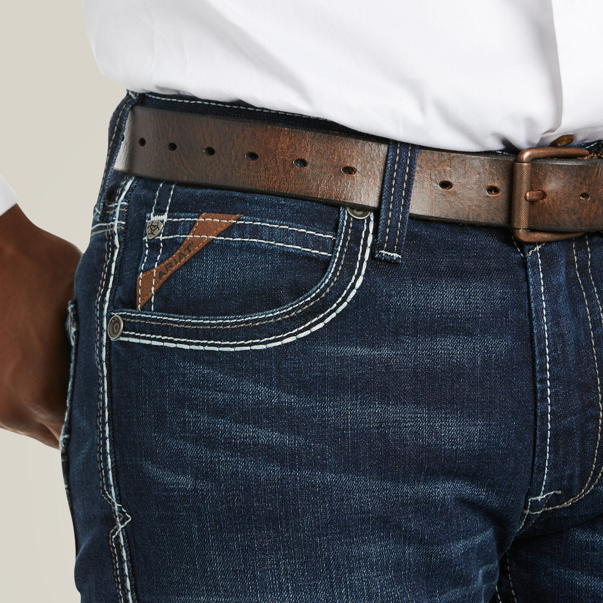 A close-up features a person in the M5 Slim Stretch Coltrane Stackable Straight Leg Jean - Nightingale, showcasing dark blue denim with white stitching and reinforced pockets. They wear a brown leather belt and a white shirt, with one hand partially visible at the side.