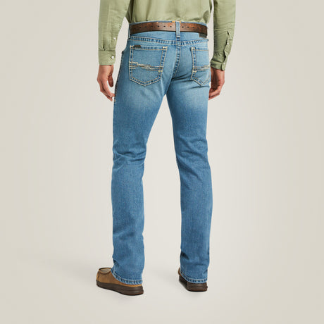 A person wears a green long-sleeve shirt, M7 Slim Stretch Julian Straight Jean - Hartwell - 10039614 with visible stitching, a brown leather belt, and brown shoes, standing like a modern cowboy with their back to the camera against a neutral background.