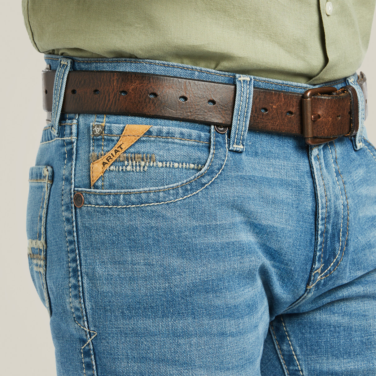 A close-up of someone wearing the M7 Slim Stretch Julian Straight Jean - Hartwell (10039614) with a brown belt and tan pocket label highlights modern cowboy style, featuring visible stitching and a green shirt neatly tucked in.