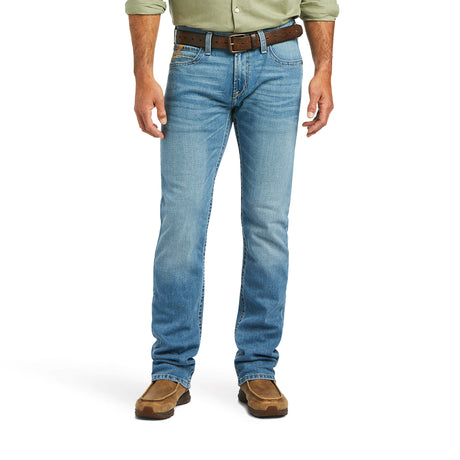 A modern cowboy exudes confidence in light blue M7 Slim Stretch Julian Straight Jeans, a brown leather belt, a green long-sleeve shirt, and tan suede shoes. Their arms rest casually at their sides against a plain white background.