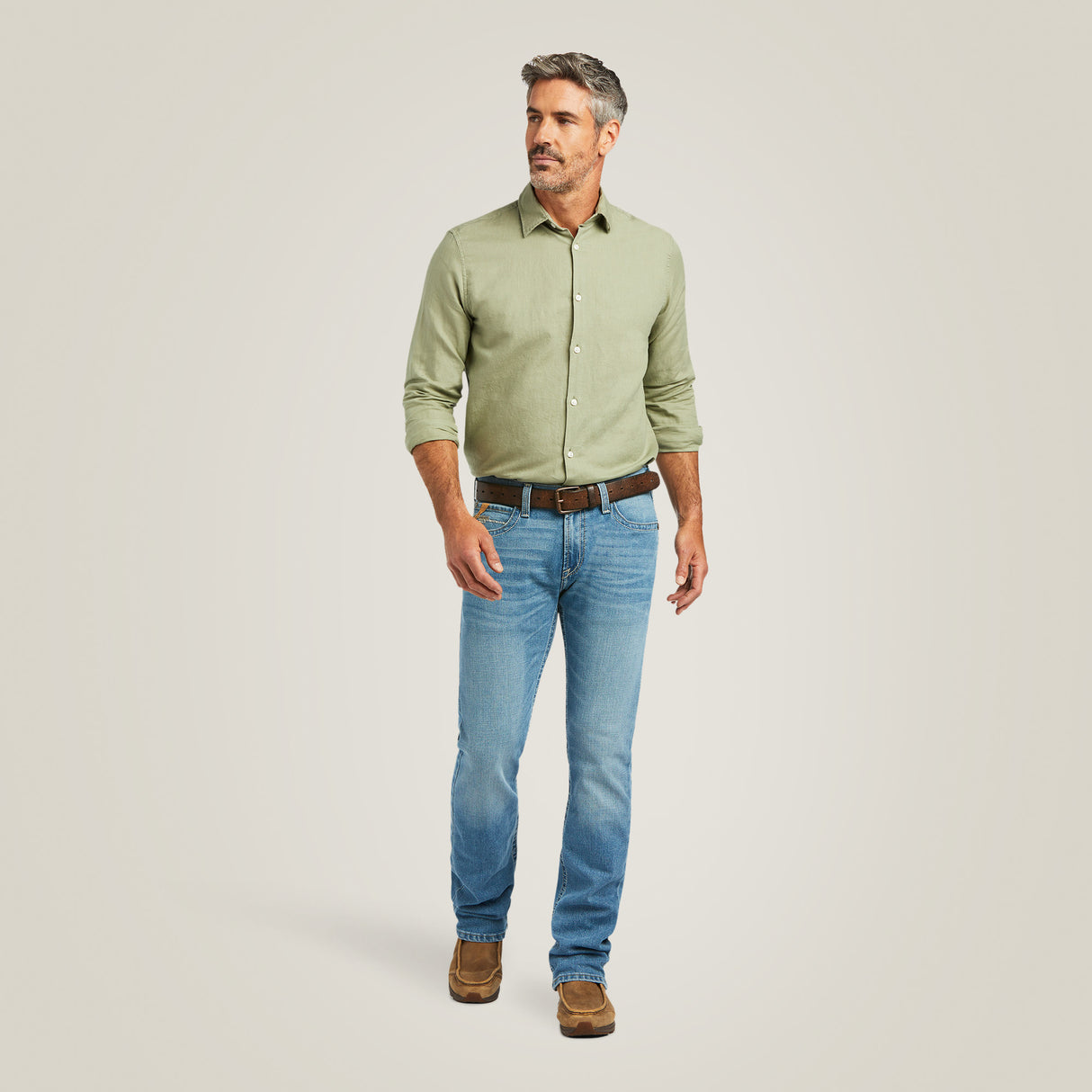 A man in a light green button-up shirt and M7 Slim Stretch Julian Straight Jeans - Hartwell stands against a neutral background, pairing his look with brown shoes. He embodies effortless style as he gazes to his right with a relaxed posture.