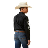 A young boy stands sideways in an Ariat Boys Black & White Team Logo Embroidered Long Sleeve Western Shirt (10045426), featuring a logo and white lettering on the sleeve and back. Hes paired it with blue jeans, a brown belt, and a wide-brimmed white cowboy hat, looking left against a plain white background.