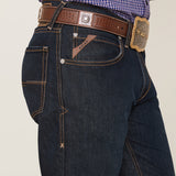 A person wears the M4 Relaxed Hansen Boot Cut Jean in Blackstone, featuring beige stitching and a brown leather belt with a decorative buckle. The jeans have a logo patch and stylized pockets, set against a plain white background.