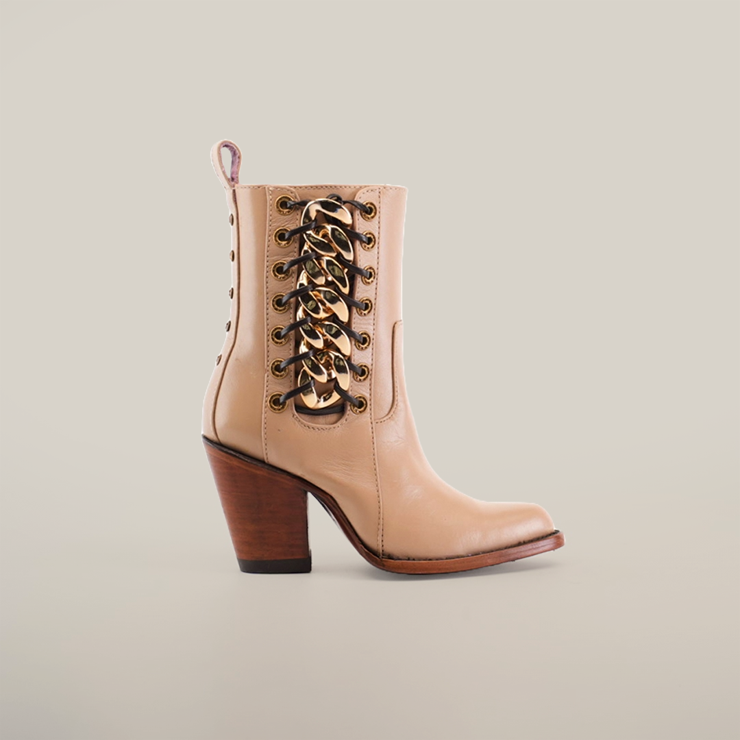 The Francia Rings Bootie Cream - J Toe, a beige leather ankle boot with a medium brown wooden heel, is shown in profile against a plain white background. It features bronze eyelets and crisscrossing lace-up details for a stylish, modern look.