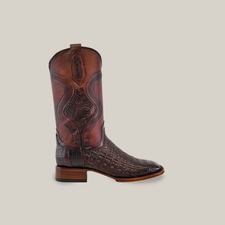 A single Exotic Caiman Belly Tail - Diamond - Tabaco - Square Toe boot stands upright against a plain beige background, featuring ornate stitching, a patterned texture, and a sturdy wooden heel, embodying exotic sophistication.